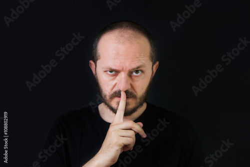  Serious man with beard and mustaches on black background in low key, making silence gesture, pst, shh 