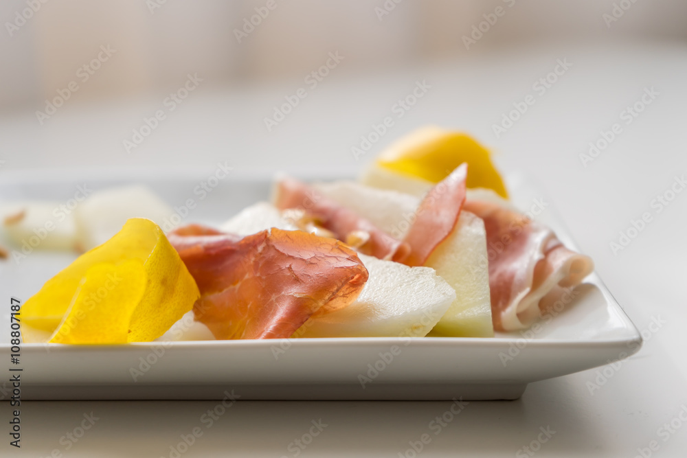 Detail of prosciutto slices with water melon. Peel and seeds. Starter in very soft focus on white plate.