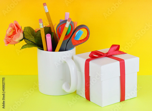 White coffee mug filled with markers, pencils, scissors and a silk rose with a gift box on a green and yellow background good for secretary's day, or administrative professionals day