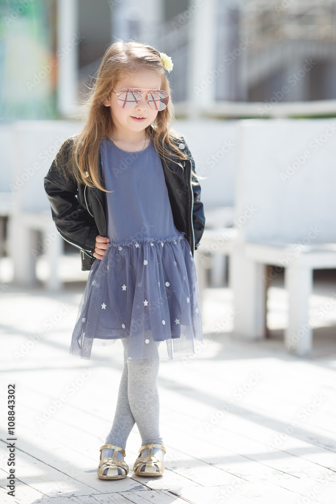 Portrait of a little girl with blond straight hair,wearing a leather jacket of black color and gray-blue gown,in her hair wears a white flower in the city posing outdoors in the spring