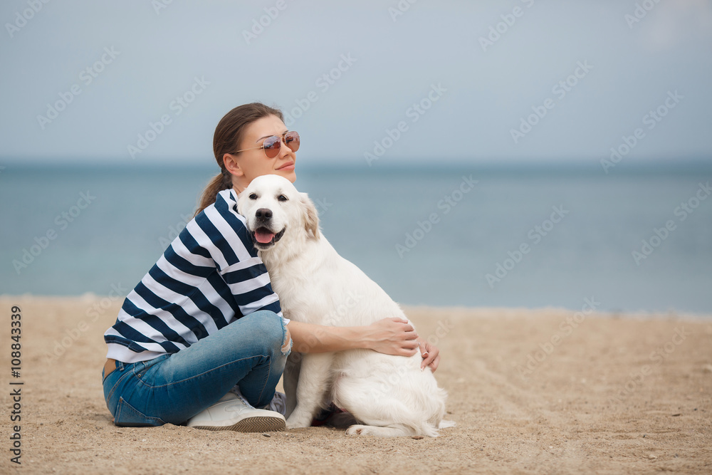 Obraz premium Portrait of young beautiful woman in dark glasses,brunette with a beautiful smile, sitting on a sandy beach against the blue of the ocean,embracing a beloved friend - a dog breed Golden Retriever
