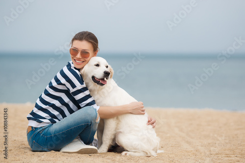 Portrait of young beautiful woman in dark glasses,brunette with a beautiful smile, sitting on a sandy beach against the blue of the ocean,embracing a beloved friend - a dog breed Golden Retriever