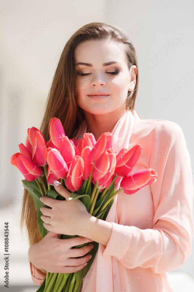Young beautiful brunette woman with long straight hair and brown eyes,dressed in a beige jacket and a white t-shirt posing in summer on the street with a bouquet of red tulips Young beautiful brunette woman with long straight hair and brown eyes,dressed in a beige jacket and a white t-shirt posing in summer on the street with a bouquet of red tulips