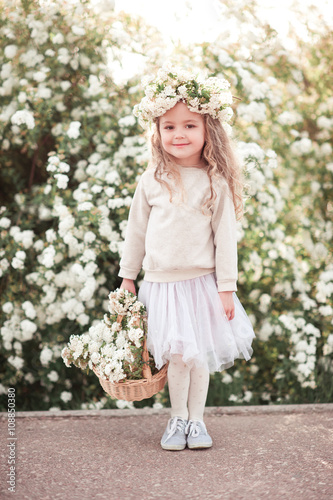 Smiling baby girl 3-4 year old holding basket with flowers outdoors. Looking at camera. Wearing stylish skirt and sweatshirt.