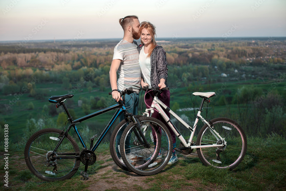 Obraz premium Beautiful young loving couple hugging at sunset, there are two bikes. The concept of happiness, peace and love