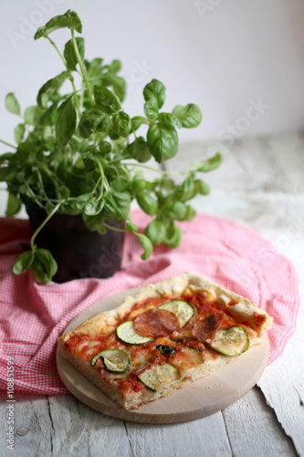 Slice of homemade pizza with tomato sauce, zucchini, spicy salami, mozzarella and basil leaves. On white rustic table, decorated with checkered tablecloth and basil pot. Selective focus.