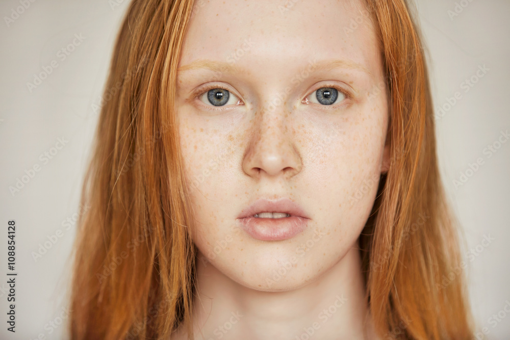Close up isolated portrait of Caucasian teenage girl with long red hair, blues eyes and healthy freckled skin looking at the camera against gray wall background. Human face expressions and emotions