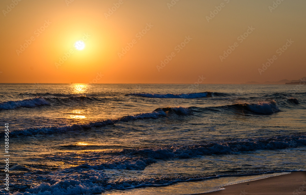 Sunrise over the sea, the rolling calm waves, sandy beach. Skay orange, waves blue.