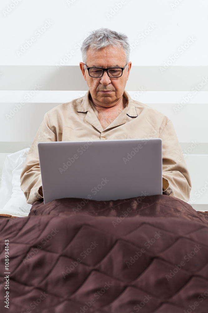 Senior man is lying in bed and using laptop