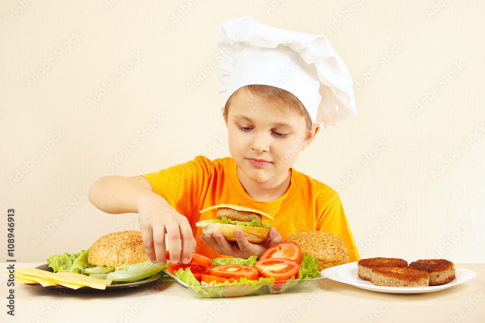 Little boy in chefs hat puts tomato on the hamburger Little boy in chefs hat puts tomato on the hamburger