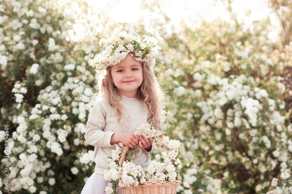 Smiling blonde kid girl holding basket with flowers outdoors. Looking at camera. Wearing floral wreath. Smiling blonde kid girl holding basket with flowers outdoors. Looking at camera. Wearing floral wreath.