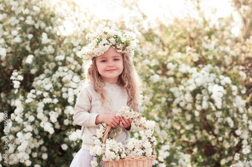 Smiling blonde kid girl holding basket with flowers outdoors. Looking at camera. Wearing floral wreath. 