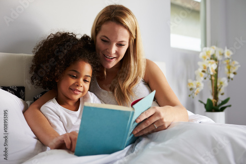 Caucasian mum and black daughter read a book together in bed