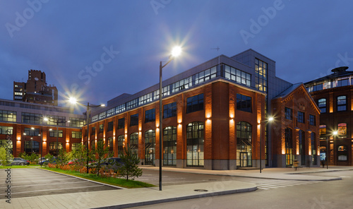 Extensive office complex exterior in loft style. Red brick buildings of former factory, gasholders. Evening architecture lighting, street lamps.