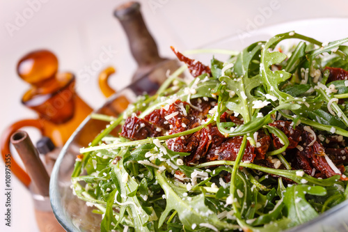 Green healthy salad with arugula and sun-dried tomatoes and parmesan cheese on a wooden kitchen table. Pepper, peppermoll, utensils for olive oil, vinegar and seasonings. Italian cuisine