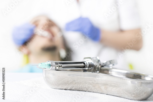 Close up of dental clinic equipment, syringe and container, with dentist and patient in the background. Dentist, stomatology and health care concept