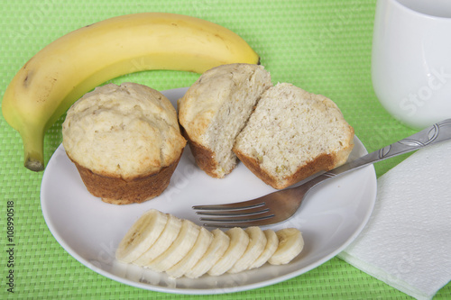 Banana bread muffins on a plate with a fork and sliced banana with napkin and mug of milk. Whole banana on place mat behind plate. Festive green placemat