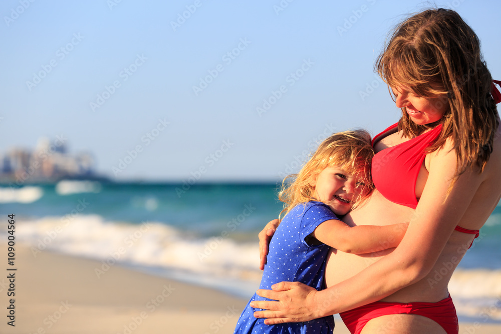 little girl hugging pregnant mother tummy at beach