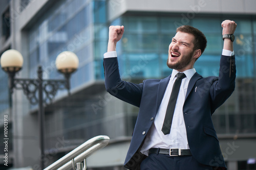 Happy businessman raising hands up against the building with glass facades © amixstudio