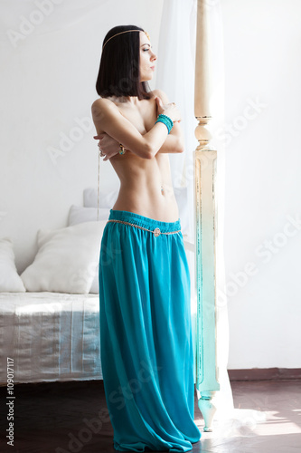 Attractive an slim woman in oriental garment and golden jewelry. She wears turquoise skirt and golden belt. She has a beautiful belly and perfect tanned skin. Bright sunny day, white room.