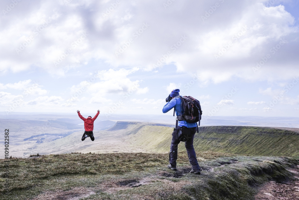 A photographer on a mountain taking a picture of a girl who is jumping on top , Pen Y Fan - obrazy, fototapety, plakaty 