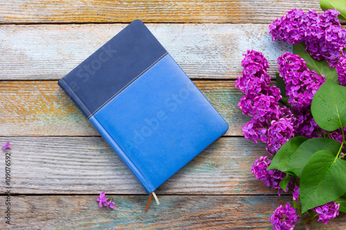 branch of purple lilac flowers with green leaves and blue notebook diary on wooden plank background