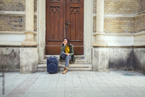 Happy young hipster girl is sitting on the stairs of the building in old town. She is holding a mobile phone and having a suitcase next to her.