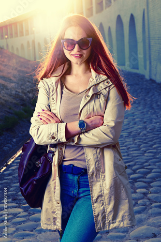 Funny stylish smiling beautiful young redhair woman in hipster clothes standing in the street with fashionable handbag,street fashion,red lips,funny sunglasses