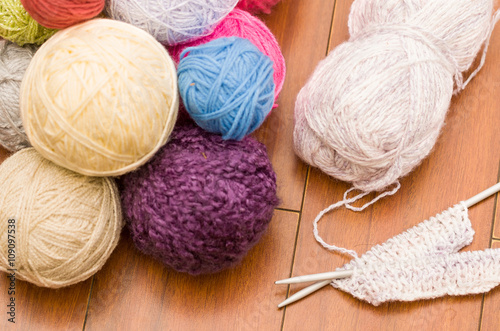 White, purple, blue and pink yarn balls piled up on wooden surface with knitting needles next to it, as seen from above