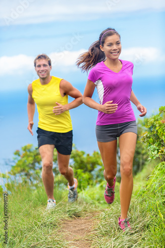 Healthy couple athletes trail running in outdoor nature path. Happy young adults training together during summer in mountains or grass park. Multi-ethnic group, Asian woman with Caucasian man trainer.