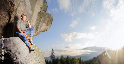 Foto Athletic male climber is sitting on the big natural boulder secured with the rope against blue sky and mountains