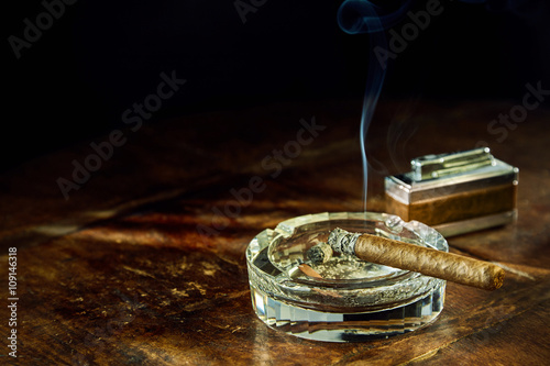 Slika na platnu Smoking cigar sitting in glass ashtray on table