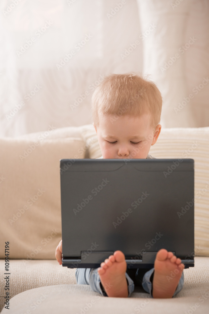 Cute little toddler boy sitting on the sofa with his laptop and looking at the window. Learning with computer. Early development. Child and computer. Cute little toddler boy sitting on the sofa with his laptop and looking at the window. Learning with computer. Early development. Child and computer.