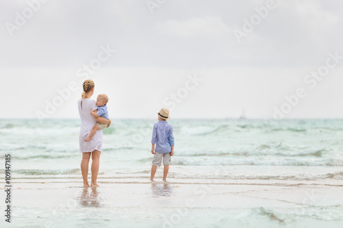 Young mother in white dress walking with her two little boys along the ocean beach. Woman with baby and boy enjoying vacation by the sea. On the empty beach. Motherhood. Water background. copy space.