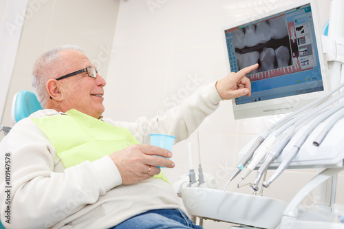 Old senior man on a dental clinic. Sitting in a chair and look X-Ray on computer