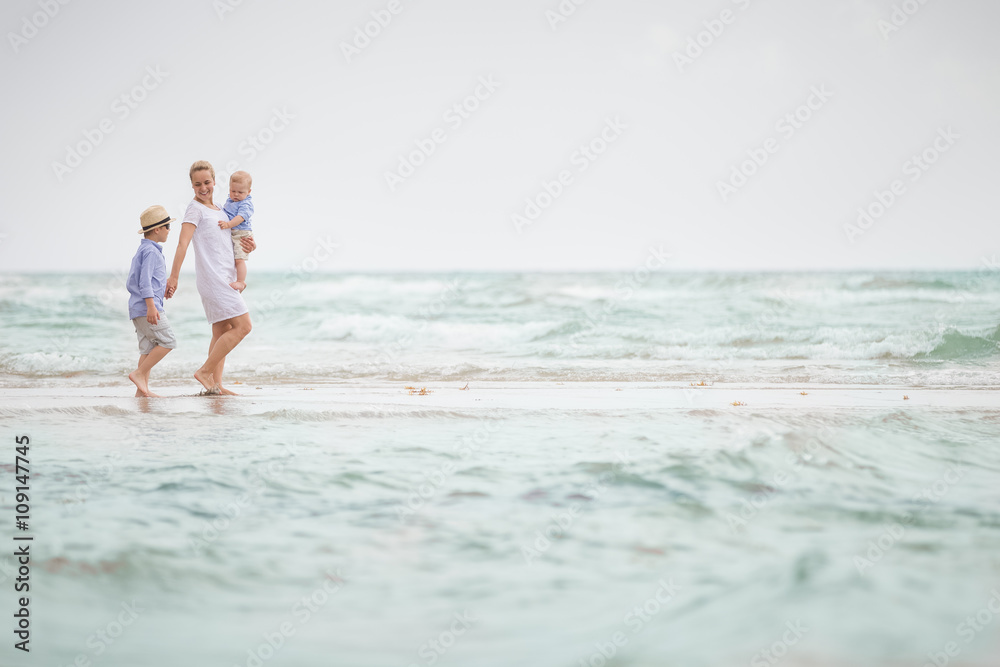 Young mother in white dress walking with her two little boys along the ocean beach. Woman with baby and boy enjoying vacation by the sea. On the empty beach. Motherhood. Water background. copy space.