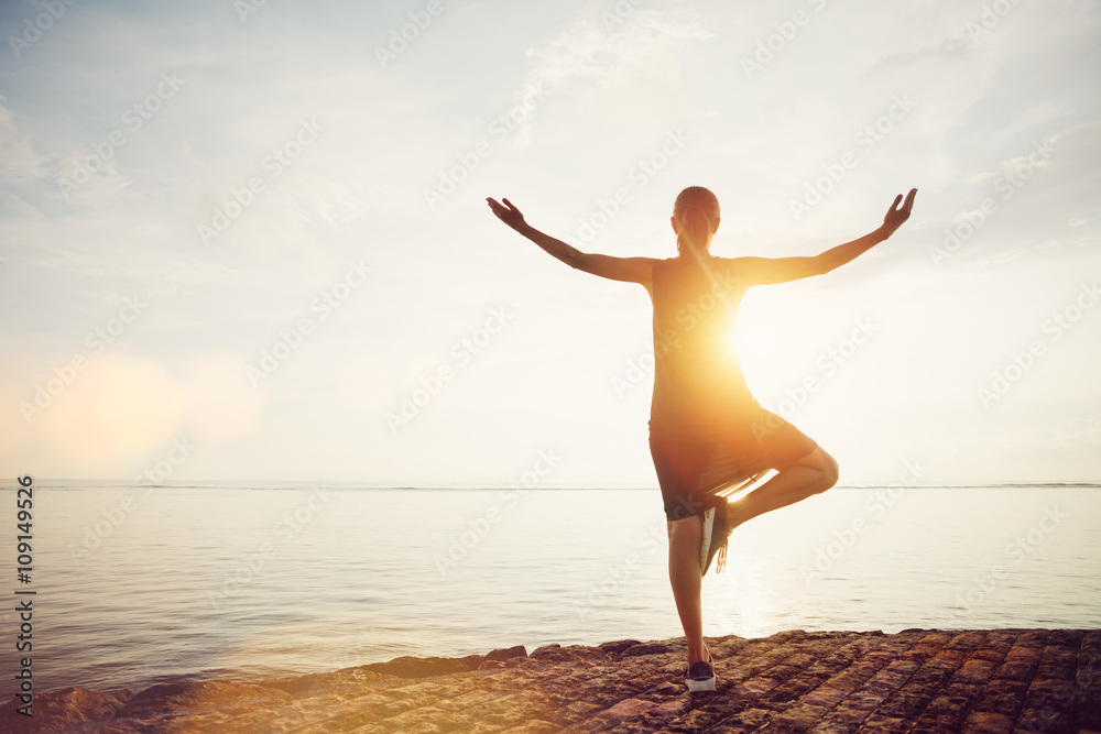 Young woman standing in yoga pose on the beach and meeting sunrise. Intentional sun glare, lens flare effect