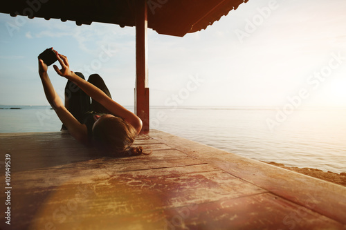 Woman touching mobile phone and resting near ocean, sun shelter on the beach. Blurry effect, lens flare effect, intentional sun glare