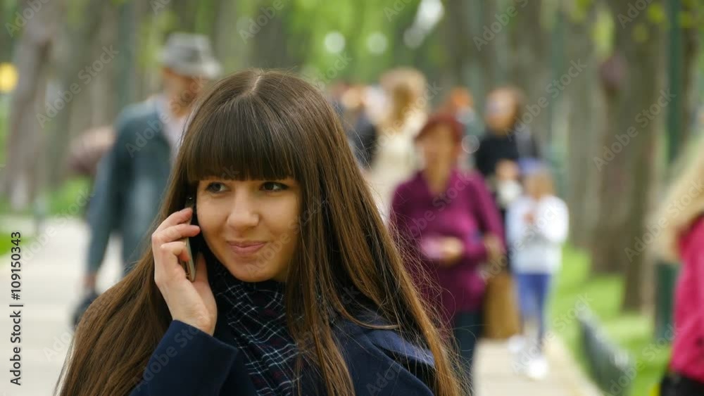 Attractive young woman in blue coat talking on the mobile phone in the alley in the park. Happy girl talking on smartphone, outdoors. Close-up Attractive young woman in blue coat talking on the mobile phone in the alley in the park. Happy girl talking on smartphone, outdoors. Close-up