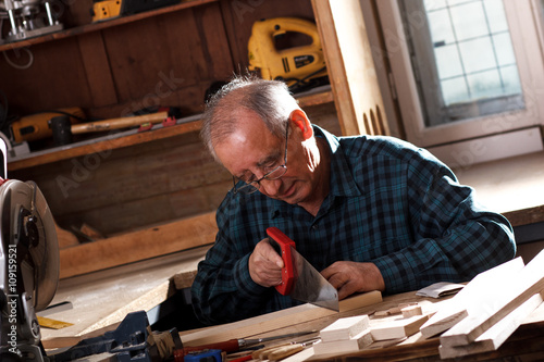 Senior carpenter working in his workshop. He is cutting wooden plank with handsaw.