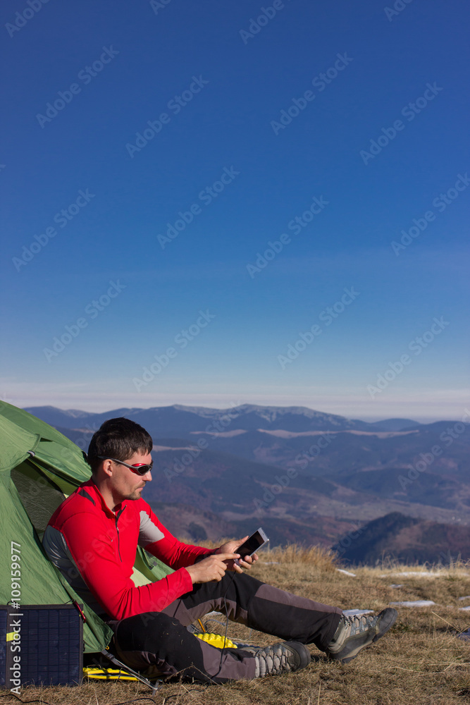 Fototapeta premium The guy in the mountains, hike with a backpack, and a solar panel for charging accumulators.