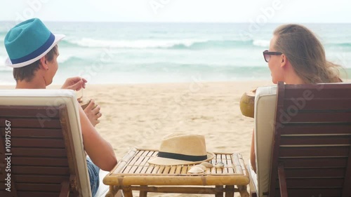 Sweet young couple in beach longers drinking fresh coconuts. Waving blue sea and white sand in the background photo