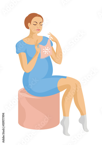 young, beautiful future mother in a blue dress sits on a padded stool and looking at a children's jacket