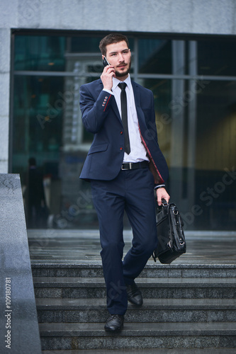 A businessman walks down the stairs and talking on mobile phone holding a briefcase against the building with a glass facade