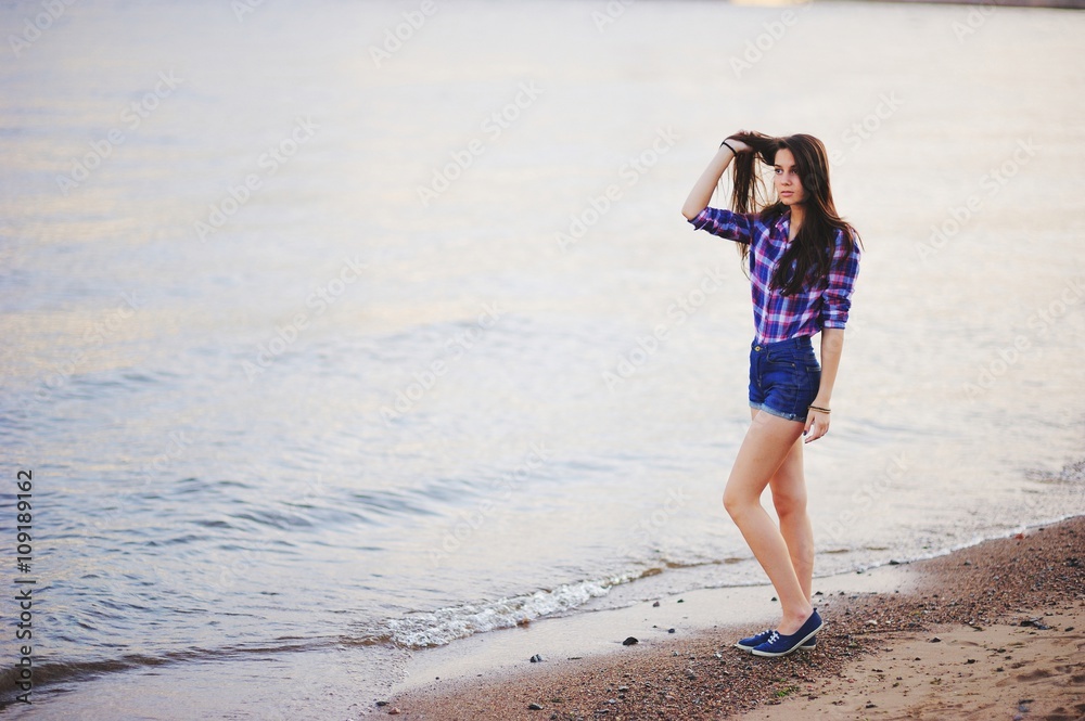 beautiful girl with long hair standing on the sand on the shore alone and sad romantic. She in shorts and a plaid shirt.