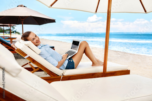 Summer Work. Portrait Of Smiling Business Woman Relaxing On Sun Lounger And Using Laptop Computer Outdoors. Beautiful Happy Girl Working Online At Beach By Sea. Communication Technology Concept. © puhhha
