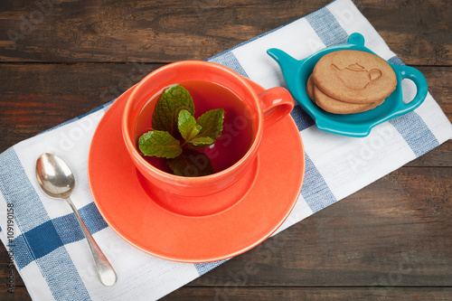 Orange teacup with tea and mint placed on teatowel together with spoon and cookies. Grungy wooden provides copy space photo