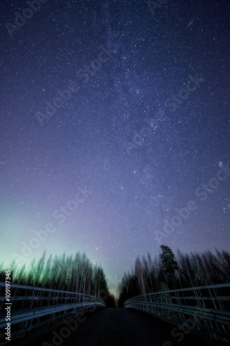 A night sky full of stars and visible milky way with a bridge on foreground. Green light of the northern lights on th left side of the sky.