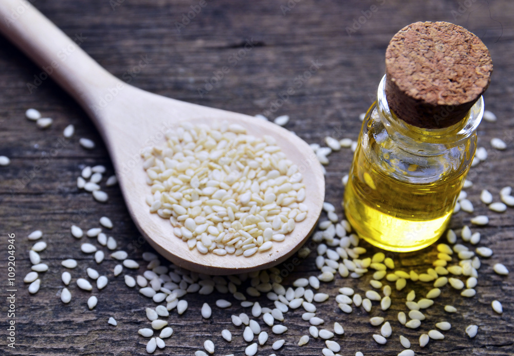 Sesame seeds on wooden spoon and sesame oil in glass jar on wooden background. Selective focus