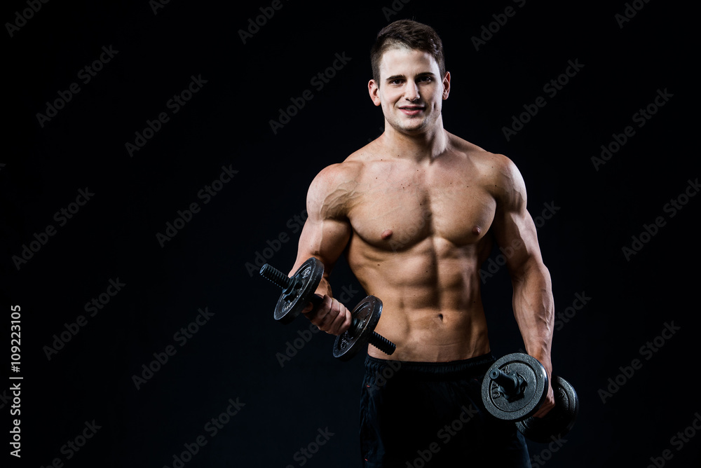 Handsome power athletic man training pumping up muscles with dumbbells in a gym. Fitness muscular body isolated on black background. Looking to the camera.
